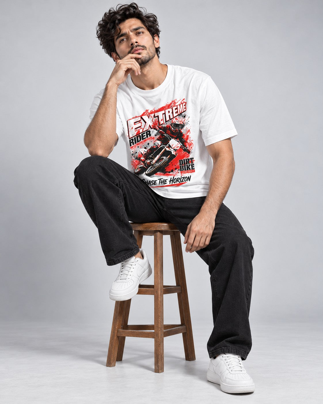 Male model wearing a white Revanx graphic printed t-shirt featuring an Extreme Rider dirt bike design, paired with black jeans and white sneakers while sitting on a wooden stool.